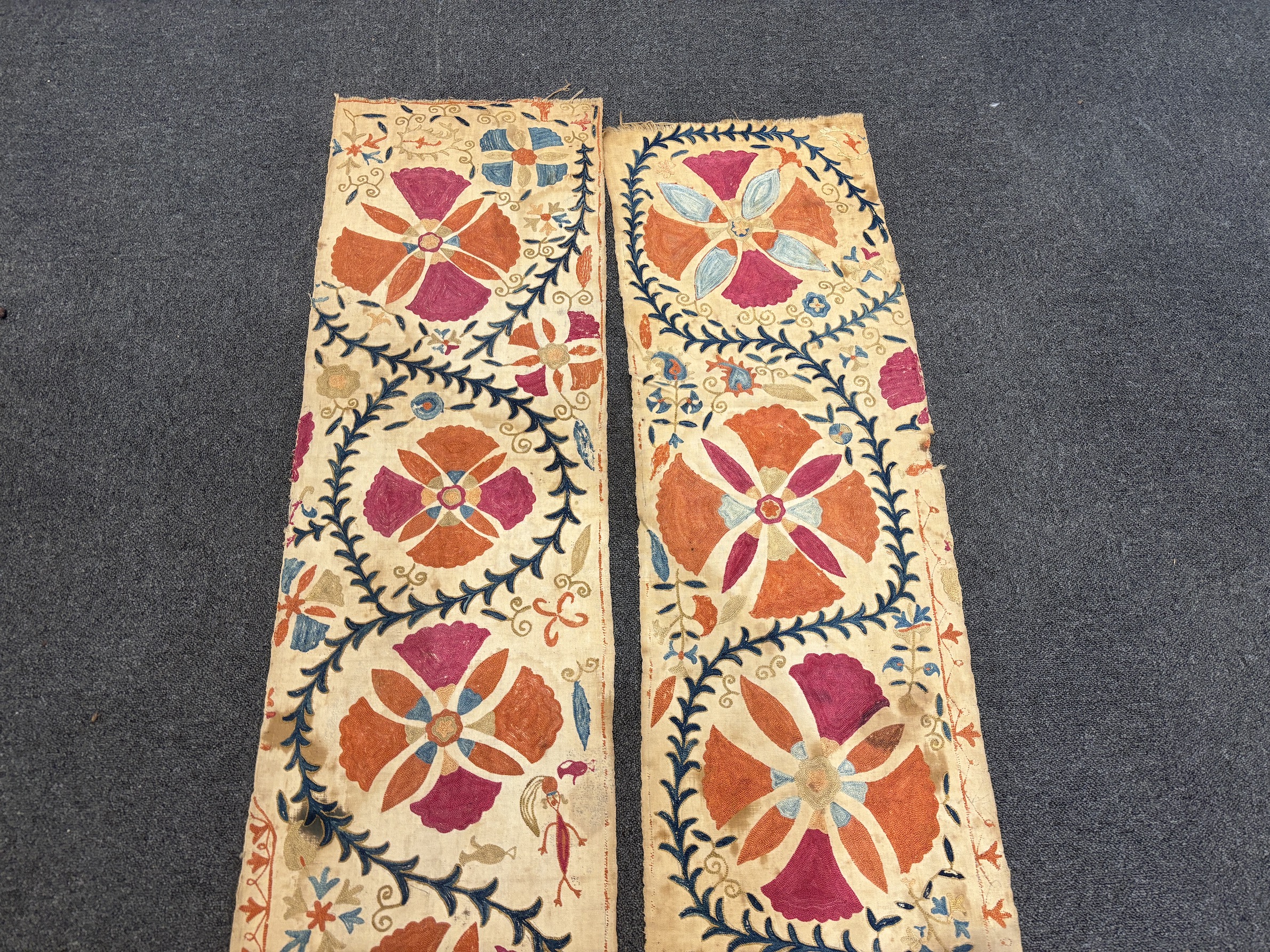 A pair of Uzbekistan long wide panels, both embroidered in mostly red, coral and blue with large symbolic flowers and trailing vines on woven cotton using mostly Burkara couching, longest 250cm long x 38cm wide
